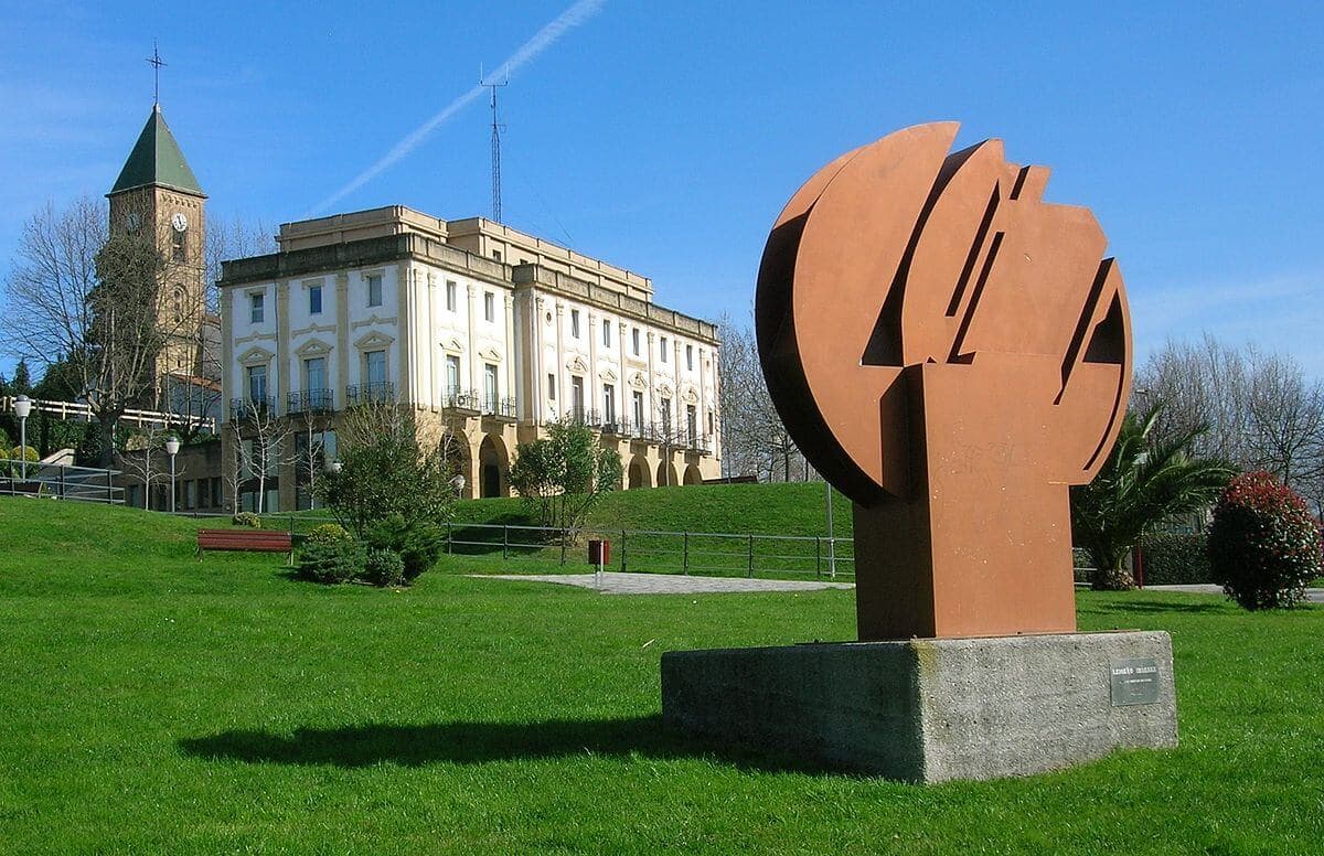 Escultura de acero corten sobre el césped frente a un edificio blanco en Leioa